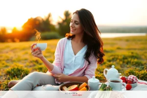 A woman is sitting on a blanket outdoors, enjoying a cup of tea. She has a light pink shirt on and her hair is down. There is a picnic table with fruit and a teapot nearby.