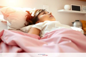 A woman lying in a hospital bed with pink blankets. She is looking up towards the camera, and there are medical monitors beside her on the bed.