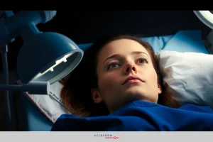 The image depicts a young woman lying down on an examination table within what appears to be a medical setting. She is wearing a hospital gown and there are medical tools, suggesting that she is receiving a medical examination or procedure.