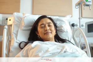 The image shows a smiling woman lying in a hospital bed. She appears to be resting or recovering and is wearing an earring. The environment suggests a clinical setting with medical equipment nearby.