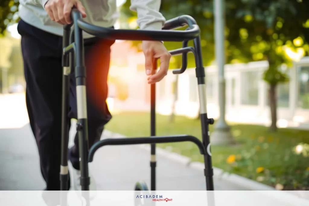 The image shows a senior adult using a walker to aid in their mobility. The person is outdoors, likely on a sidewalk, with greenery around, suggesting an urban setting with some vegetation nearby.